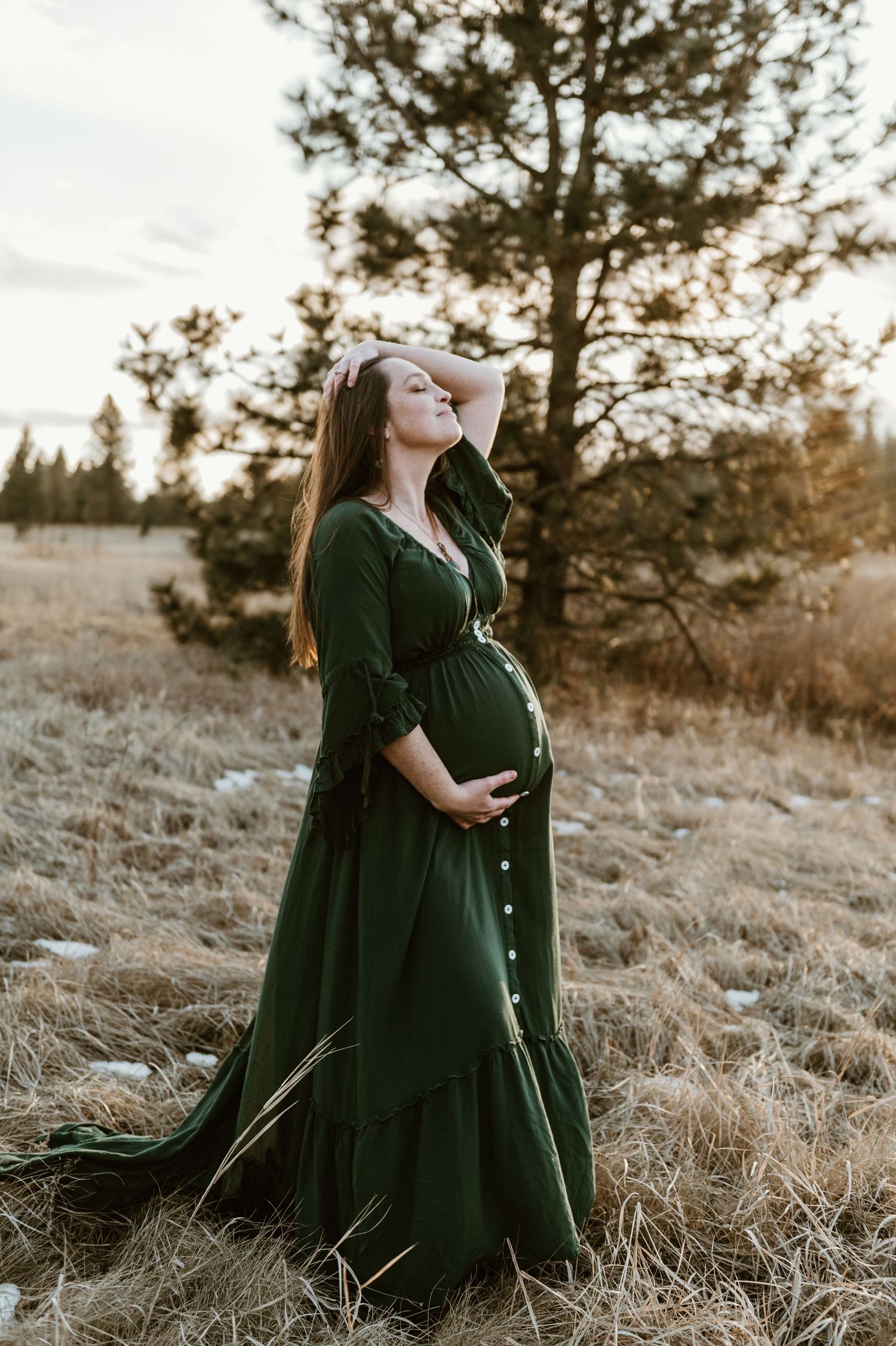 A pregnant woman in a flowing emerald dress poses outdoors at sunset with tall pine trees in the background.