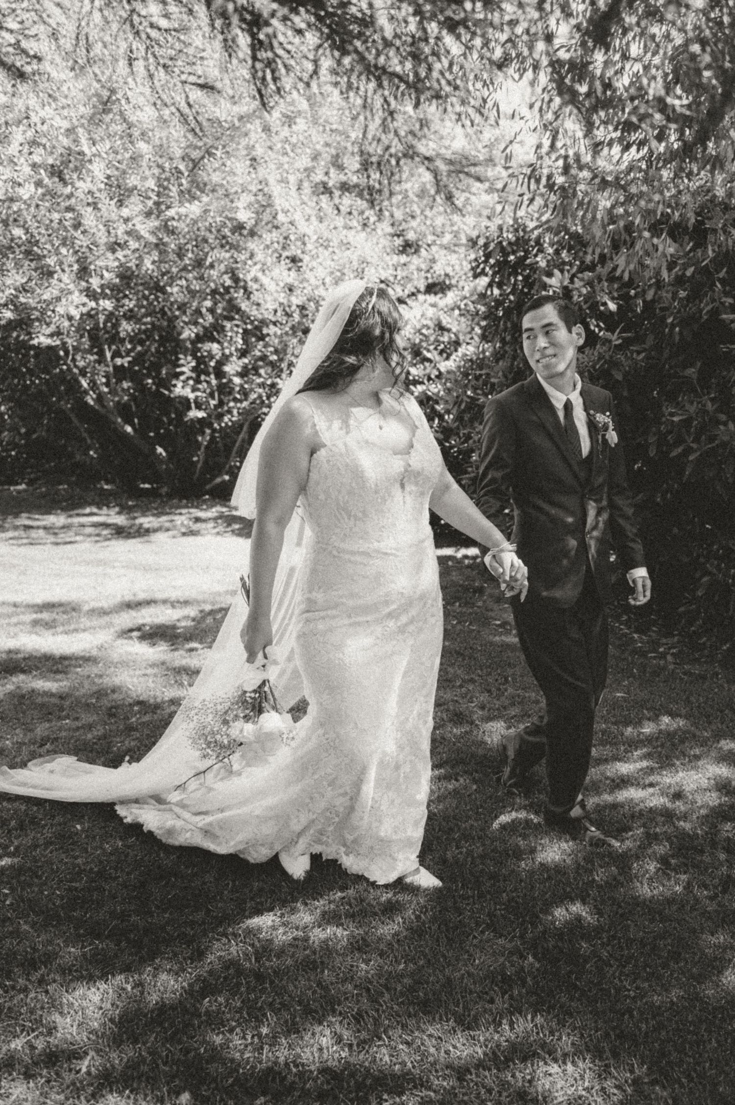 A romantic black and white wedding photo of a couple walking outdoors with the bride in a lace gown and flowing veil.