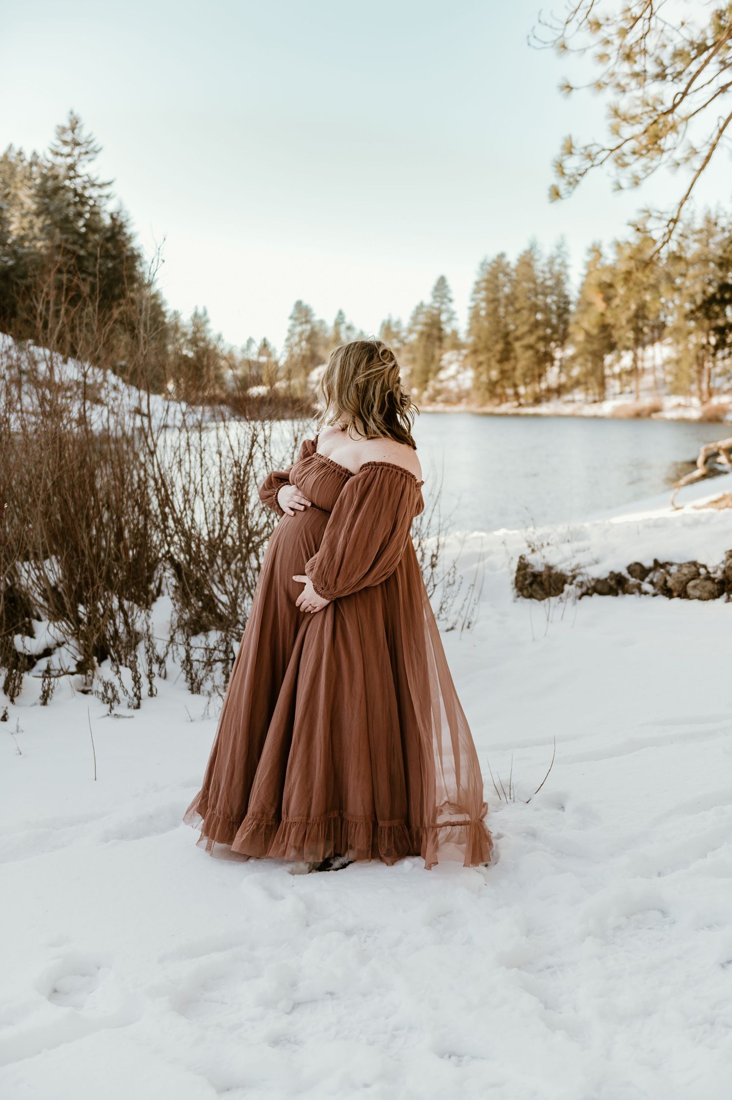 Someone in a flowing rust-colored gown stands near a snowy lake with evergreen trees in the winter landscape.
