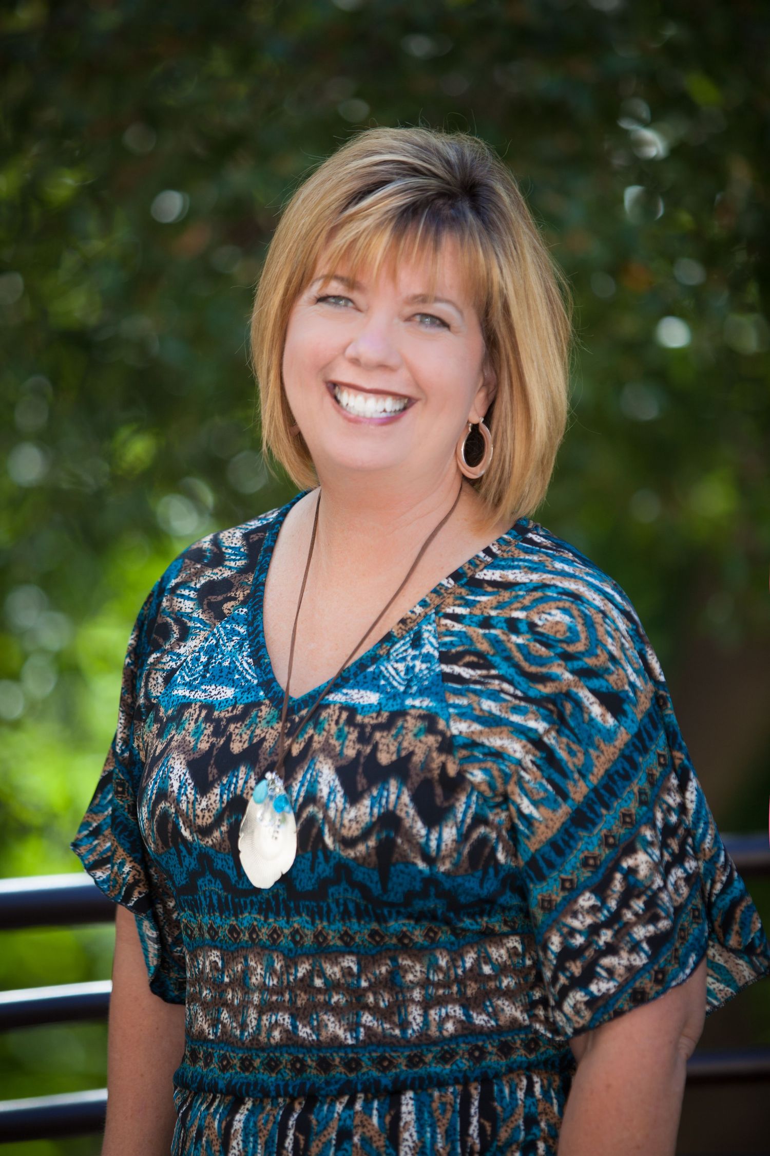 Portrait against green foliage background wearing a patterned blue top with a pendant necklace.