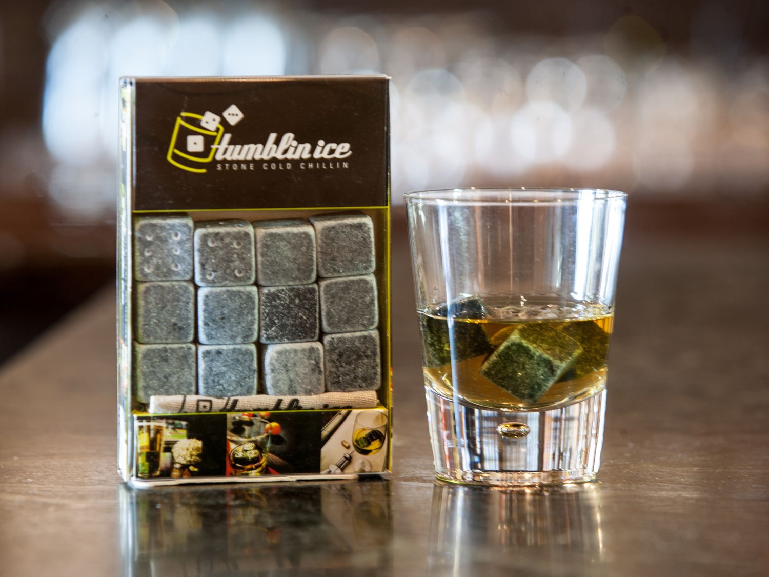Whiskey glass with ice cubes and branded packaging on dark wooden surface.