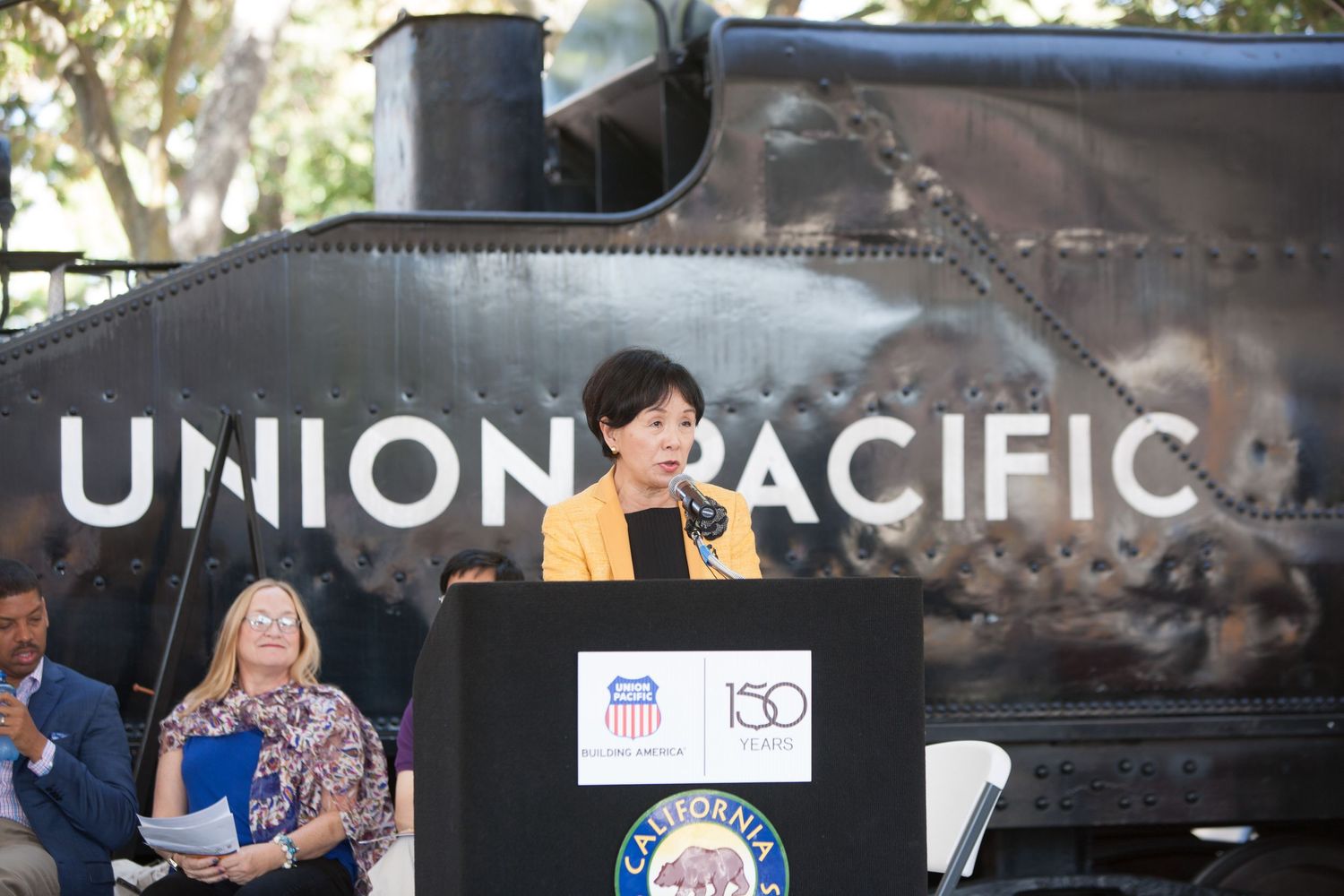 Speaker at podium in front of Union Pacific train during outdoor event.