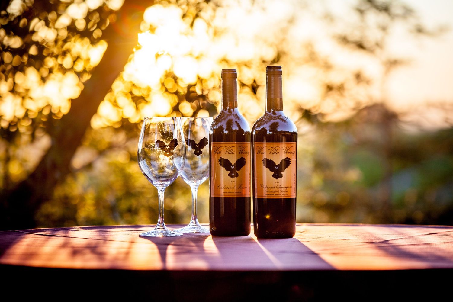 Wine bottles and glasses arranged on a wooden surface at sunset with golden bokeh lighting in the background.