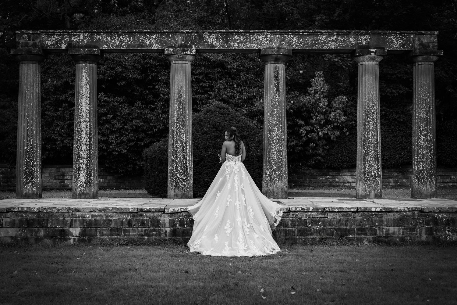 Black and white photo of a bride in a flowing ball gown standing with her back to the camera before stone columns and a leafy garden.