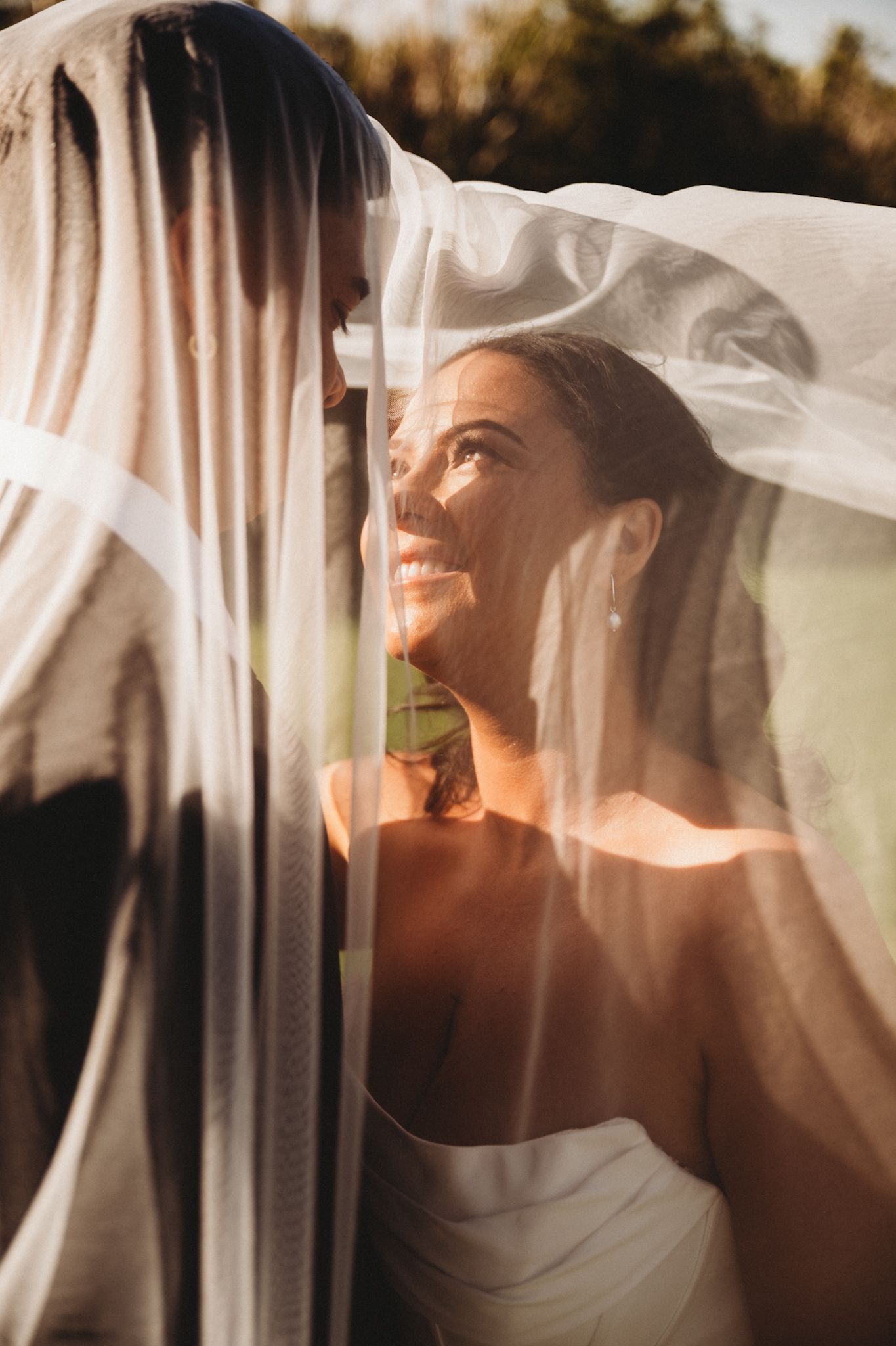 Bride smiling under veil with groom in golden sunset light, romantic outdoor wedding portrait.