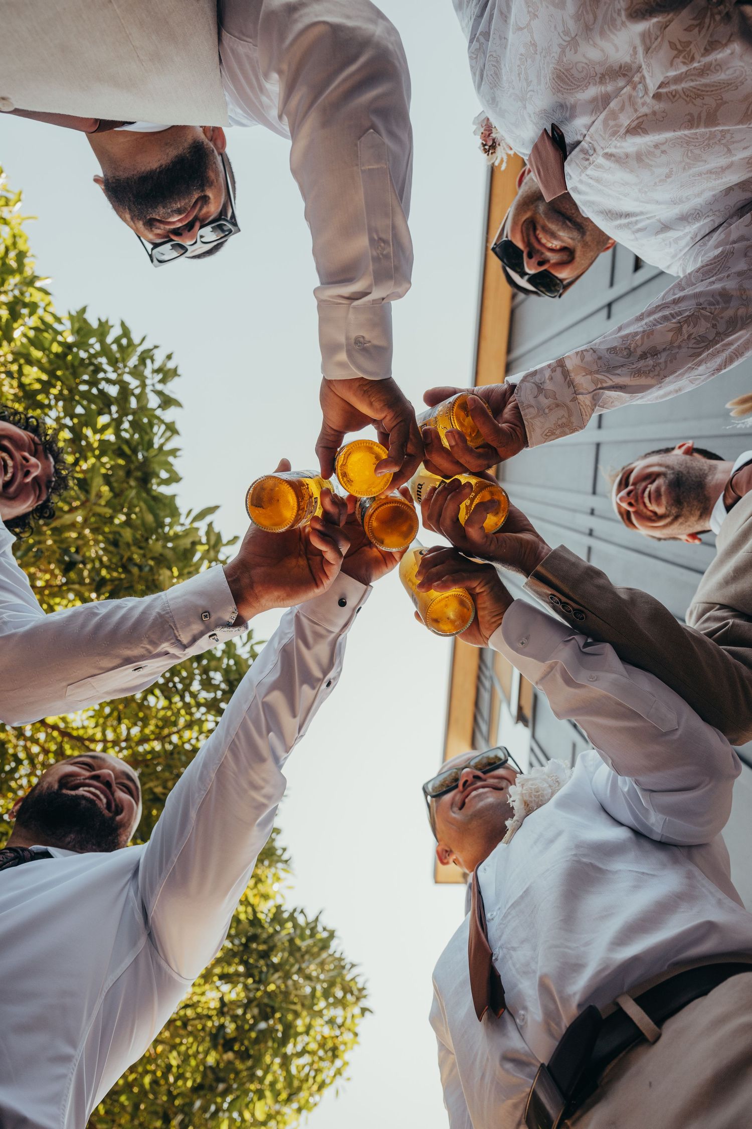 Group of men in white shirts toasting with beer bottles, photographed from below against a blue sky.