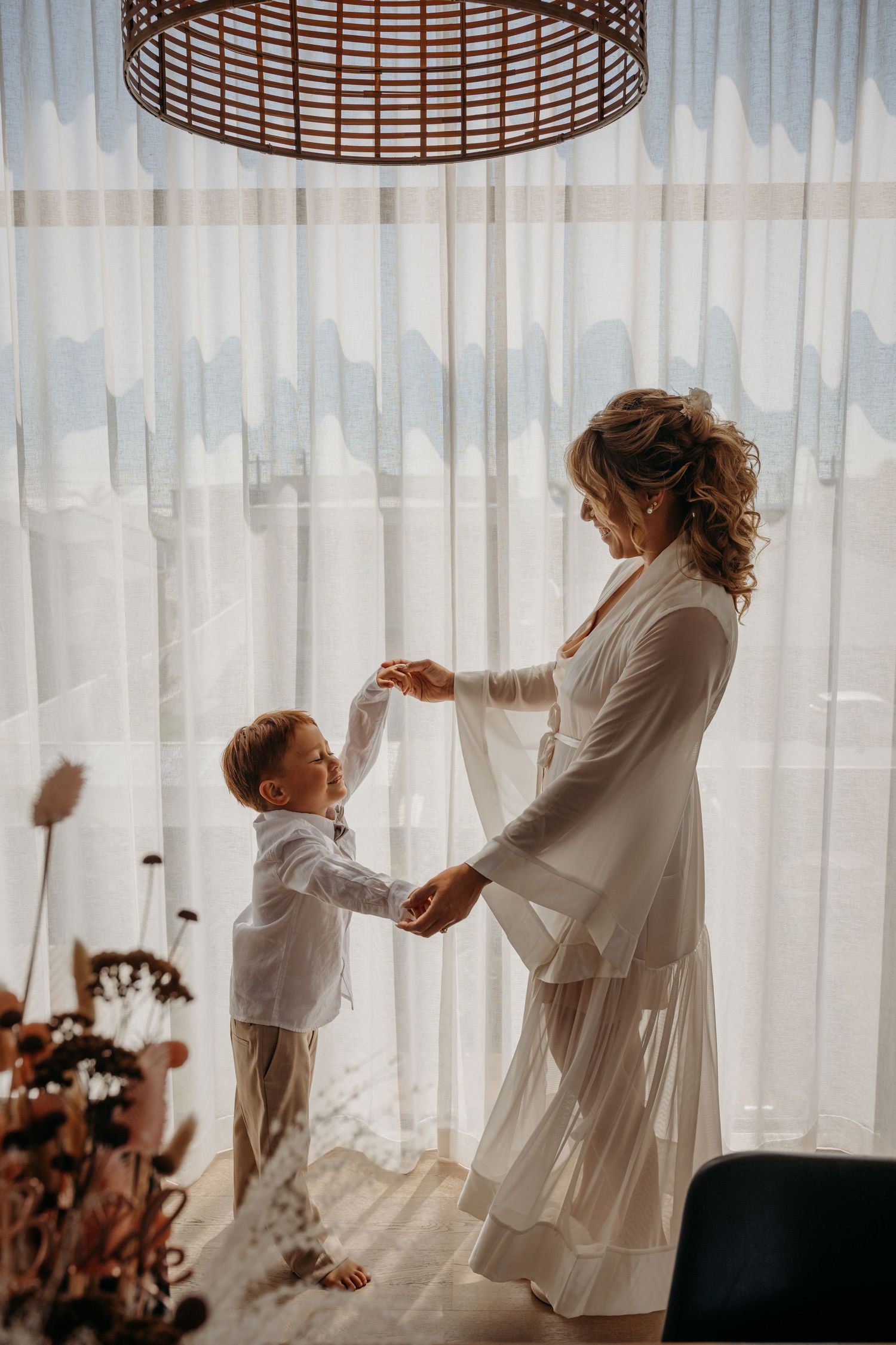 A bride in a white robe dances with a young boy in a white shirt near sheer curtains in a bright room.