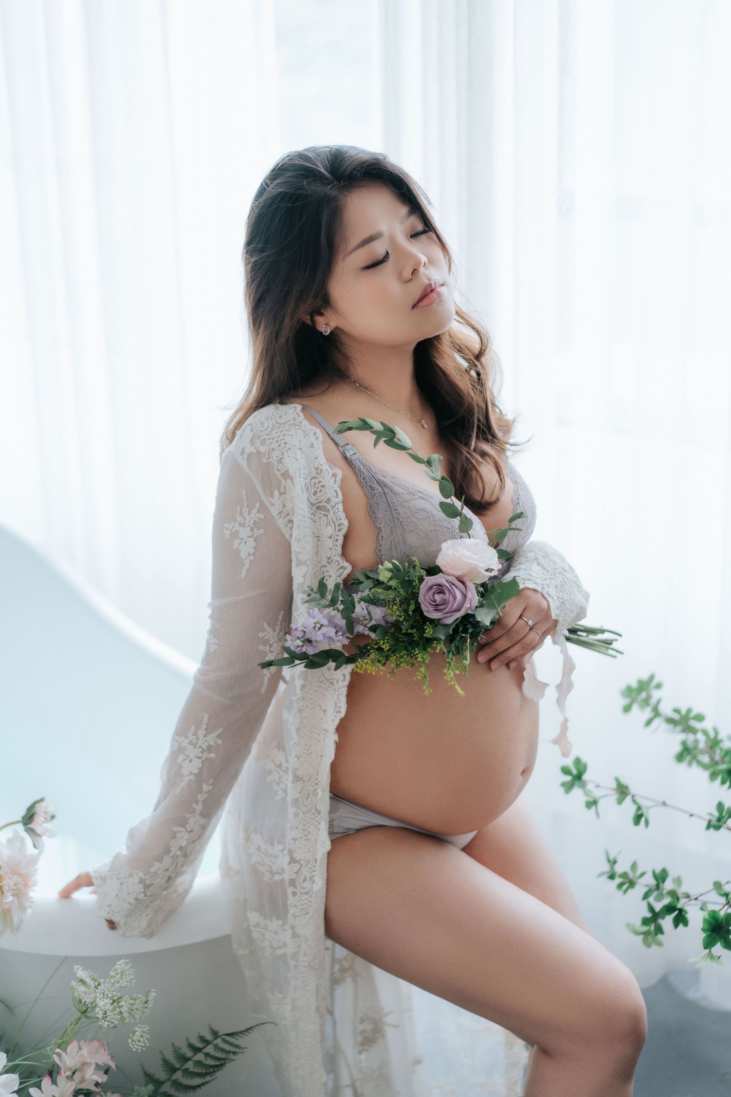 Artistic maternity photo featuring white lace robe and pink roses in soft window light.在柔和的窗光下，身穿白色蕾絲睡袍、點綴著粉紅色玫瑰的藝術孕婦照。
