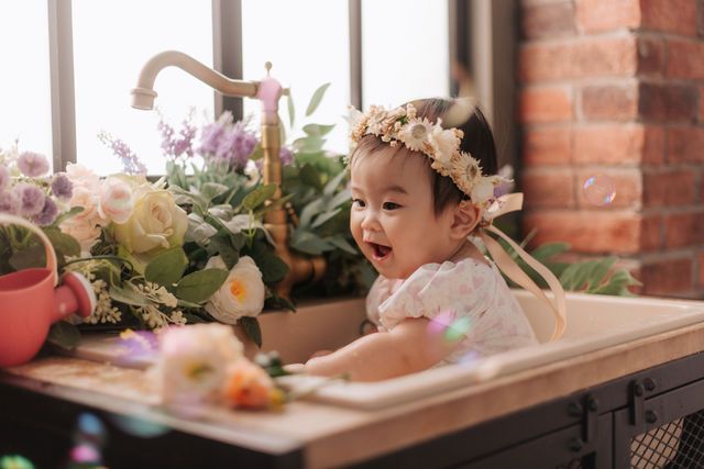 Happy baby girl in floral crown sitting in vintage sink surrounded by colorful flowers and bubbles.