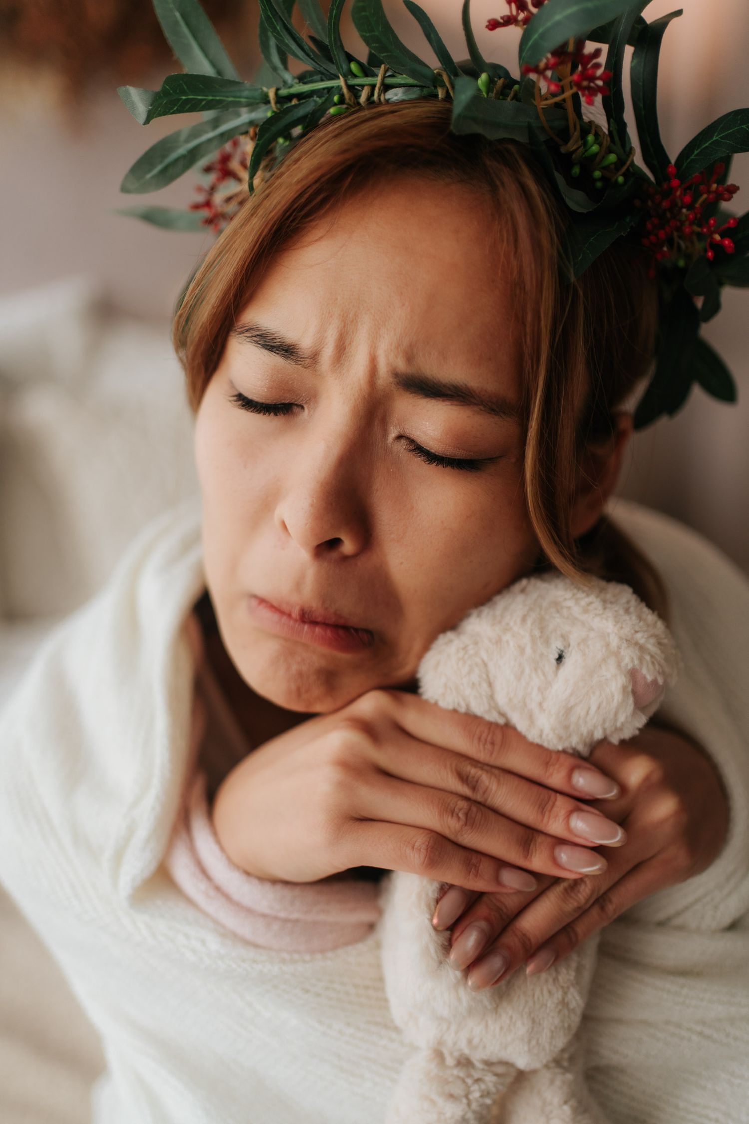 A sad woman wearing a floral crown hugs a white stuffed animal toy while sitting in a cozy white setting.