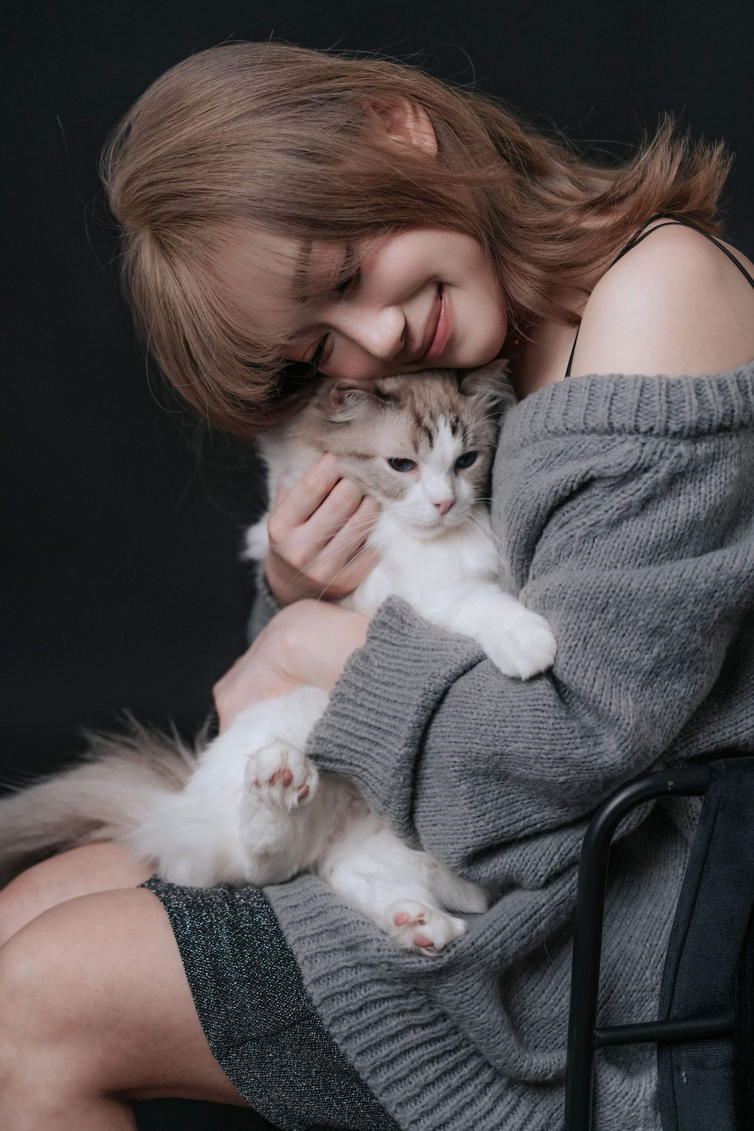 Smiling young woman in gray off-shoulder sweater cuddling a fluffy white and brown kitten against dark background.一位微笑的年輕女子身穿灰色露肩毛衣，在深色背景下