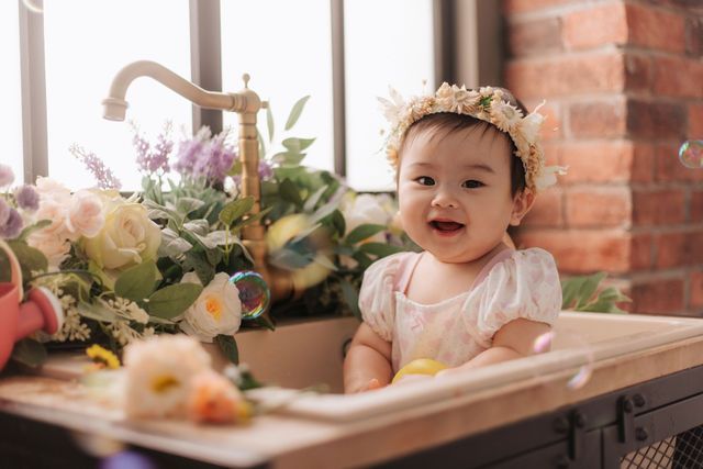 Smiling baby girl in a floral crown sitting in a decorative sink surrounded by colorful flowers.