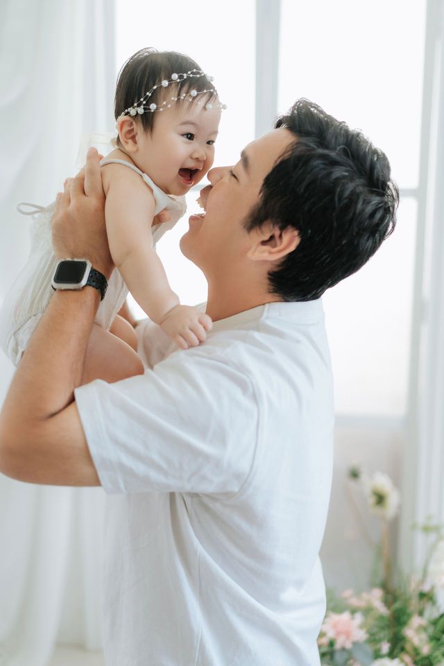 A father joyfully lifts his smiling baby girl wearing a floral headband in a bright white room.