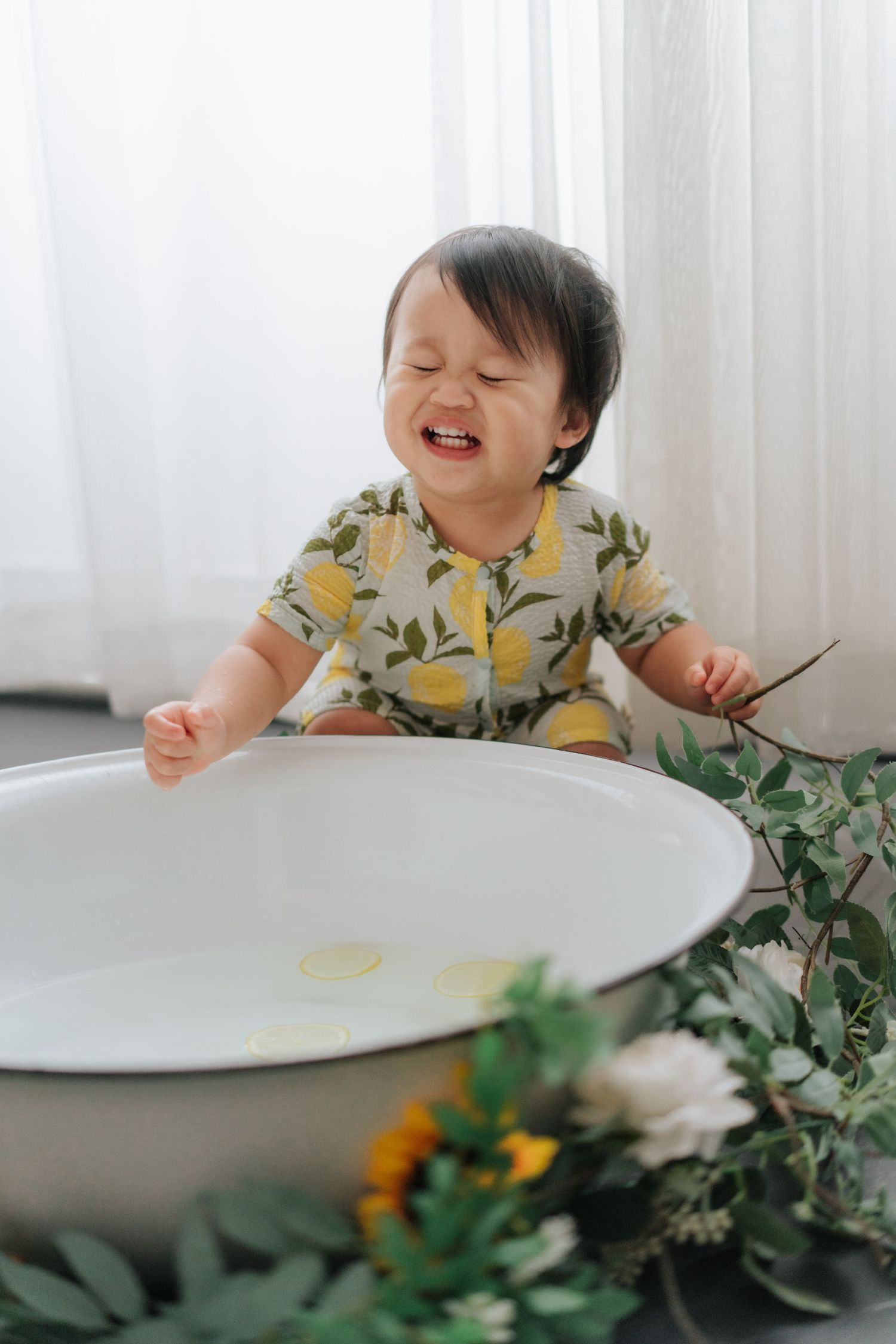 Smiling baby in lemon-print outfit playing near a white basin with flowers and greenery.