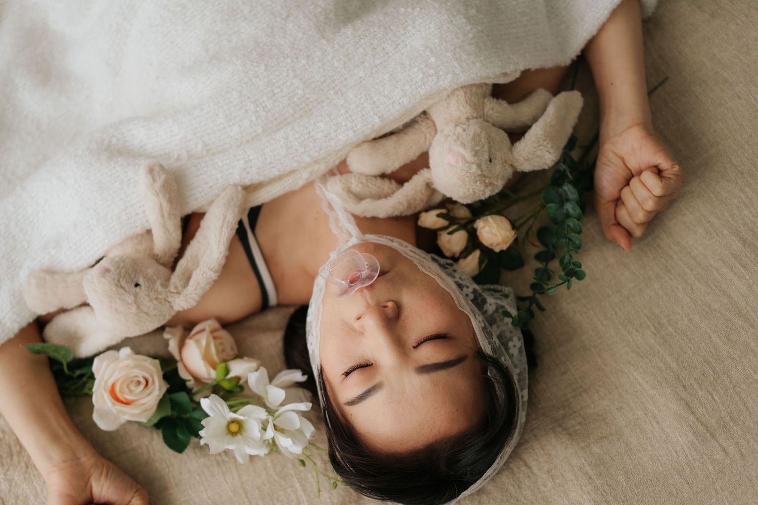 Baby sleeping peacefully surrounded by soft bunny toys and white flowers on a cozy blanket.