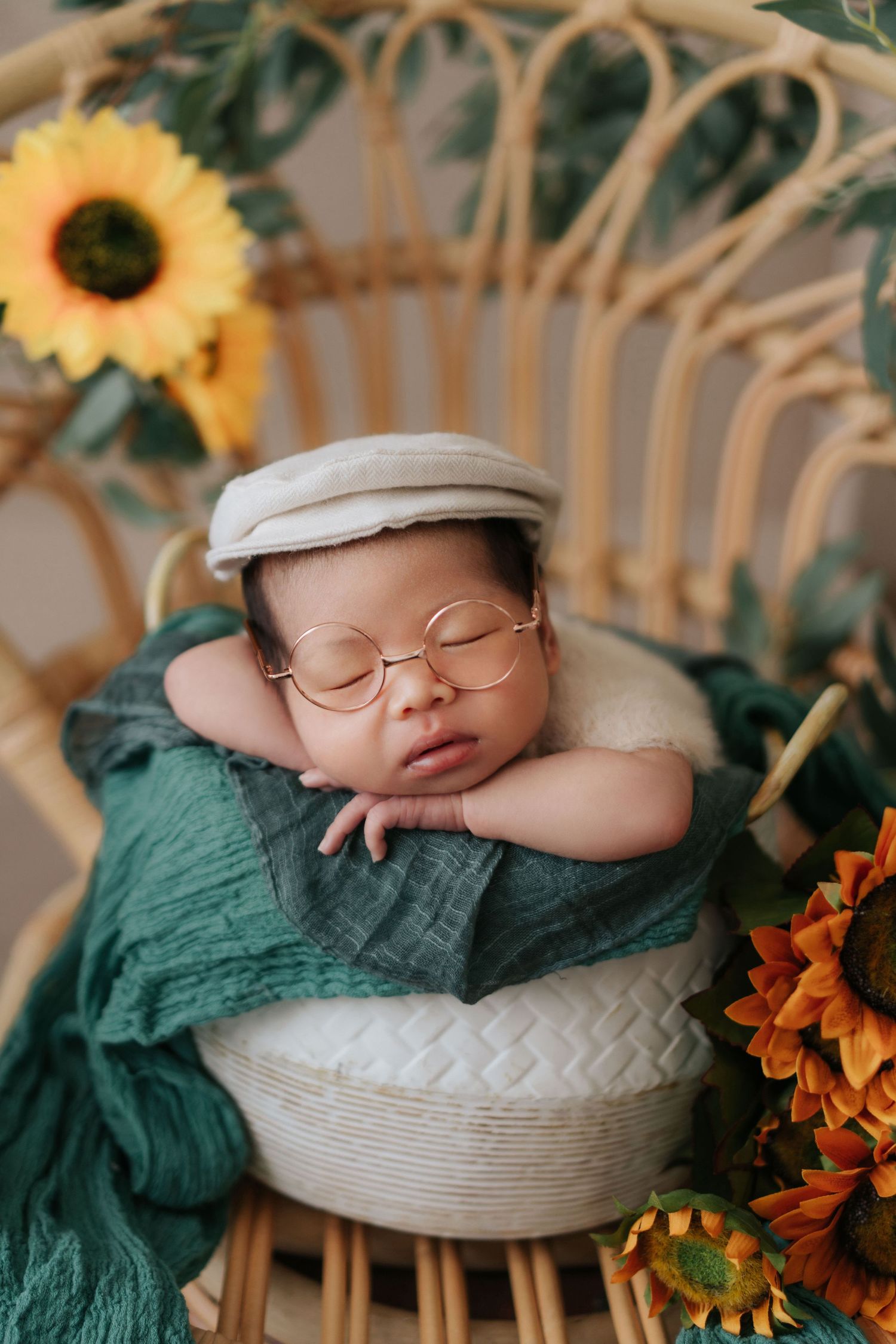 Sleeping baby in glasses and newsboy cap rests in wicker chair surrounded by sunflowers and green fabric.
