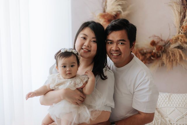 Happy Asian family portrait with smiling parents holding their baby girl in a bright, cozy white studio setting.