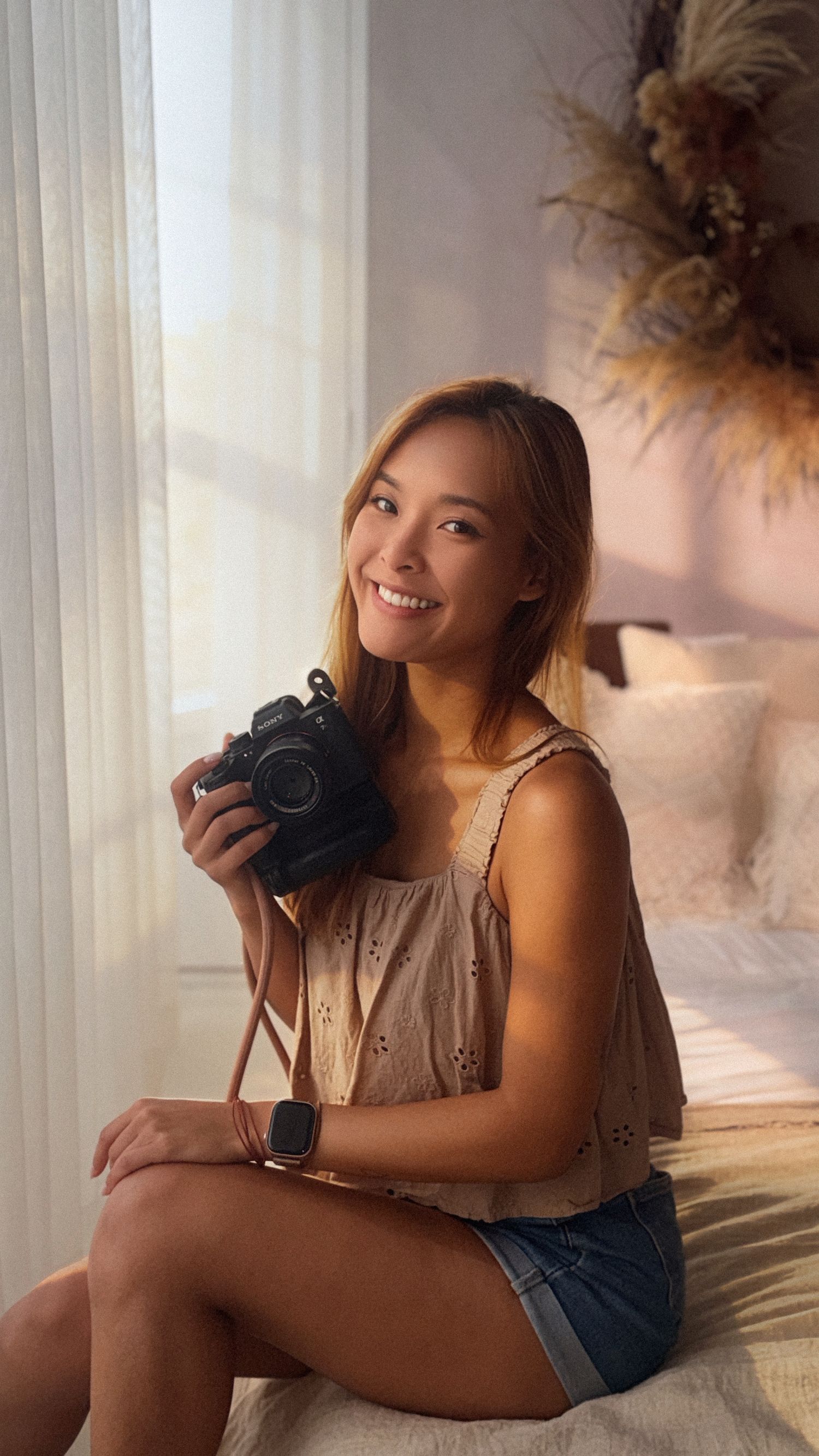 Smiling young woman holding a black camera, wearing a beige crop top and denim shorts, sitting by a sunlit window.