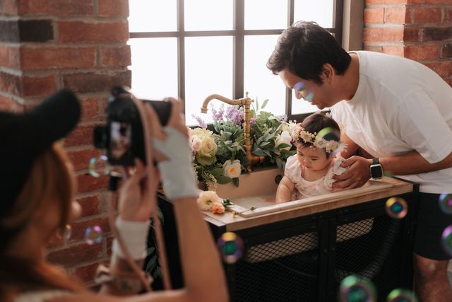 A father leans over his baby girl at a table while a photographer captures the moment, with bubbles floating in the foreground.
