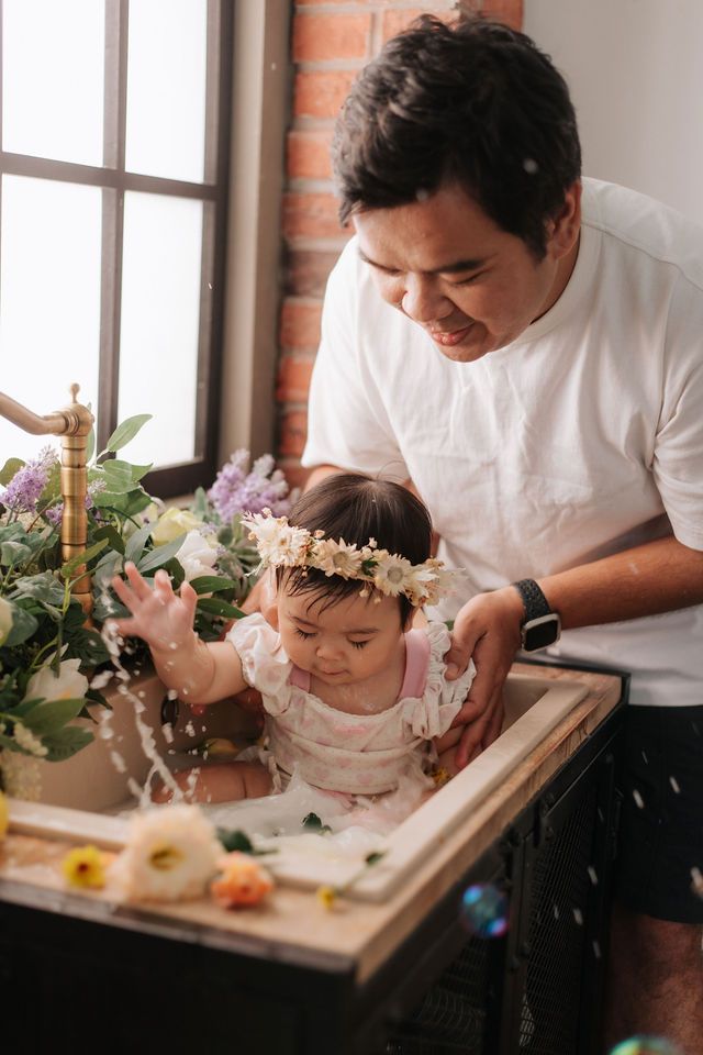 Father smiling while bathing baby girl wearing floral crown in sink decorated with flowers and greenery.