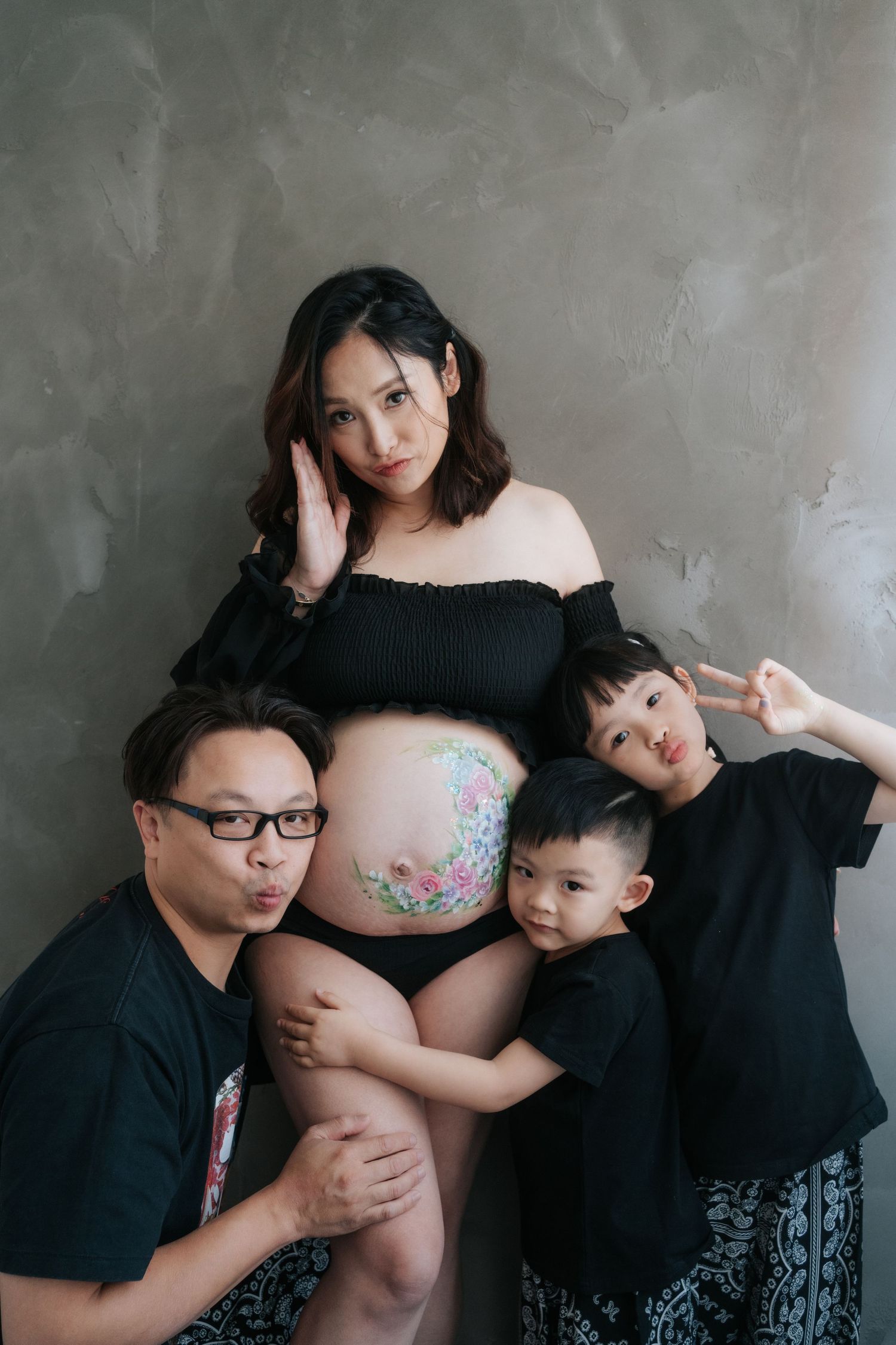 Pregnant Asian woman with floral belly paint poses with husband and two young children against gray wall.一位懷孕的亞洲女子，腹部繪有花朵圖案，與丈夫和兩個年幼的孩子在灰色牆壁