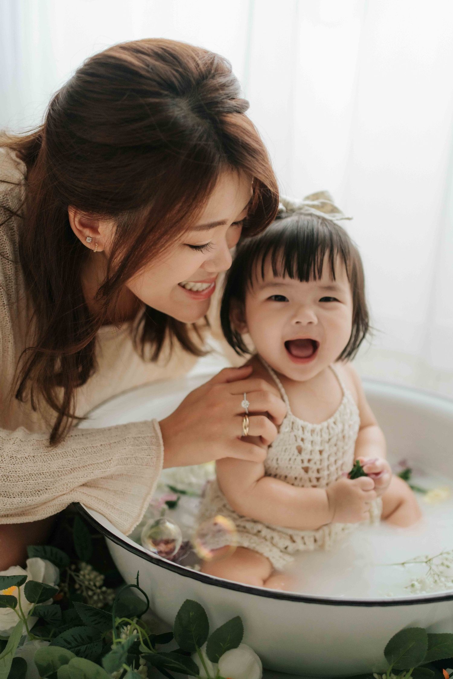 A smiling Asian mother leans over her laughing baby girl sitting in a floral milk bath tub.