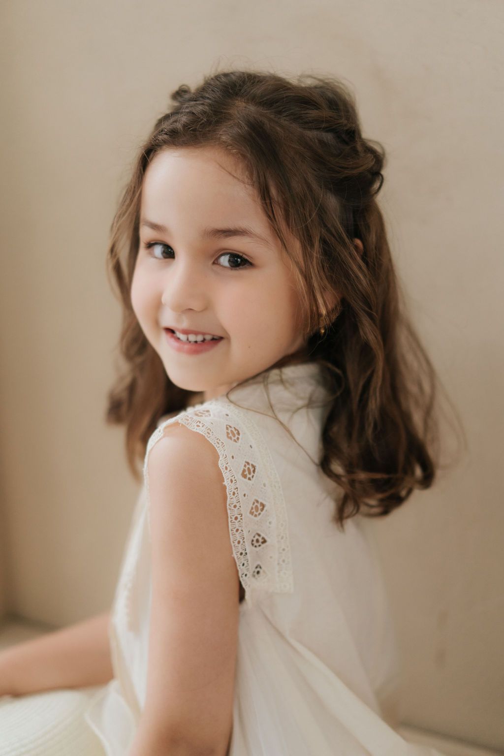 A young girl in a white lace dress smiles over her shoulder, with brown wavy hair and gold earrings.