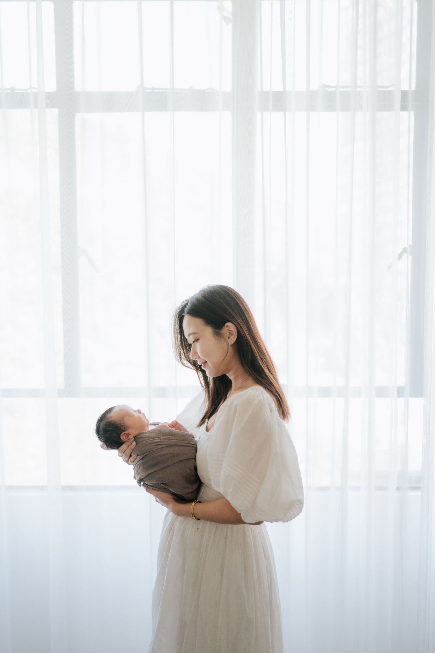 A tender moment between parent and newborn baby in a bright, sunlit room with white curtains.