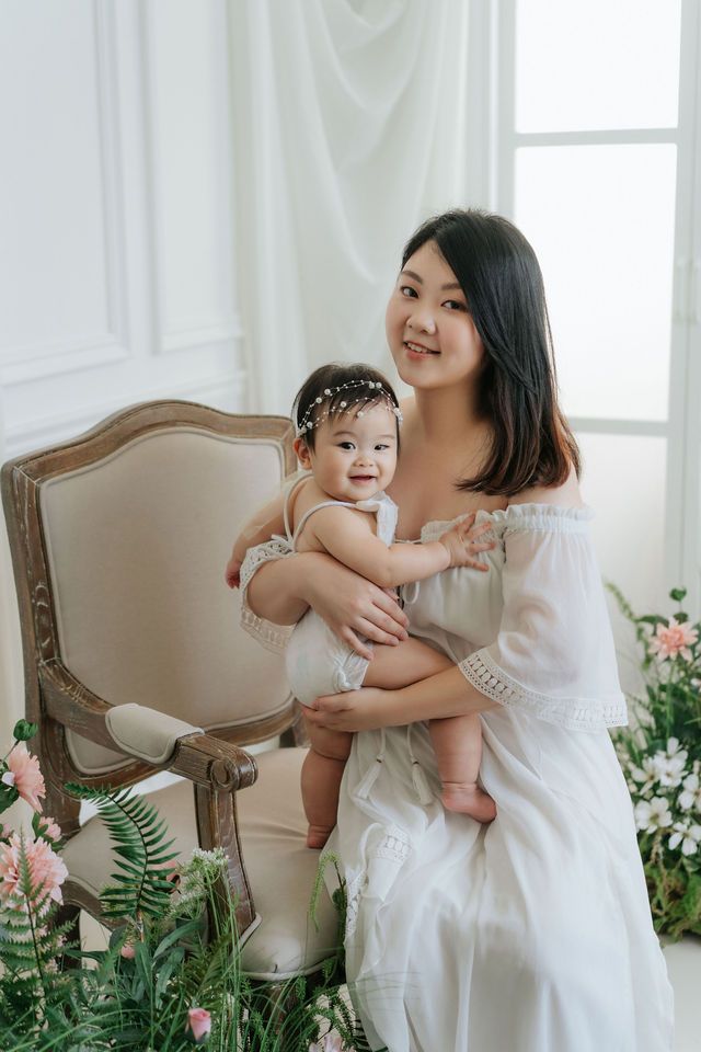 A smiling Asian mother in a white dress holds her adorable baby girl surrounded by pink flowers.