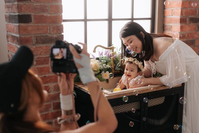 Photographer capturing a mother in white dress with baby wearing flower crown at decorated desk near brick wall.