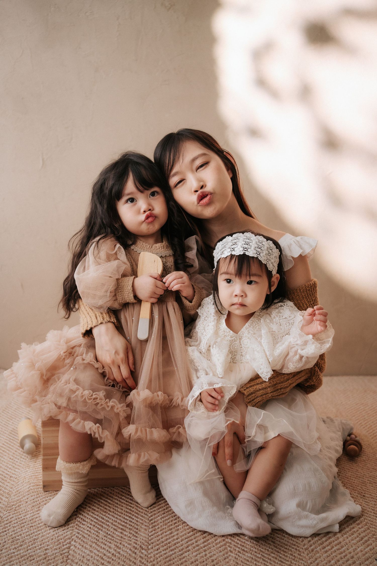 Three young Asian girls in elegant tulle dresses pose together, with the oldest making a kissing face in a soft studio setting.