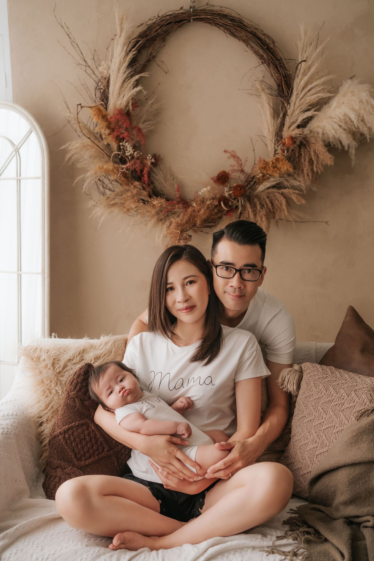 Family sits together on bed beneath boho wreath with pampas grass and dried flowers in neutral tones.