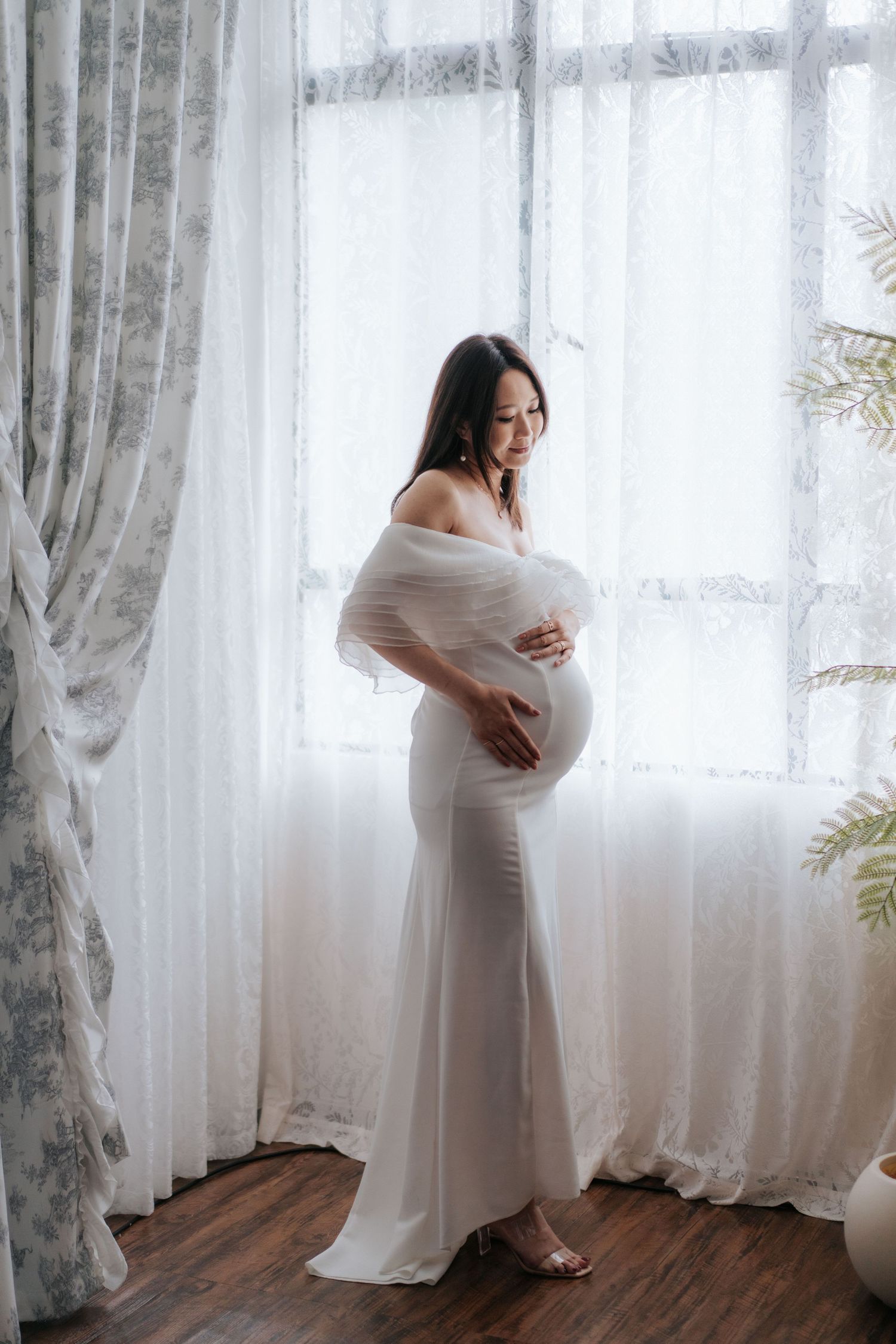 Ethereal maternity photoshoot featuring a woman in white dress by sheer curtained windows in natural light.