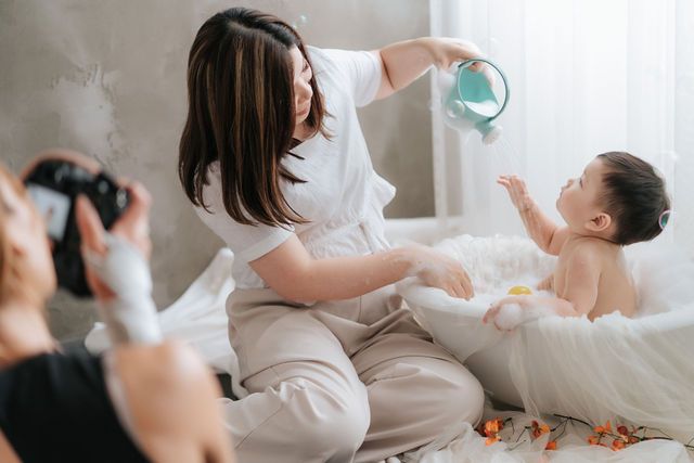 A woman bathes a baby on a bed while a photographer captures the moment with a camera.