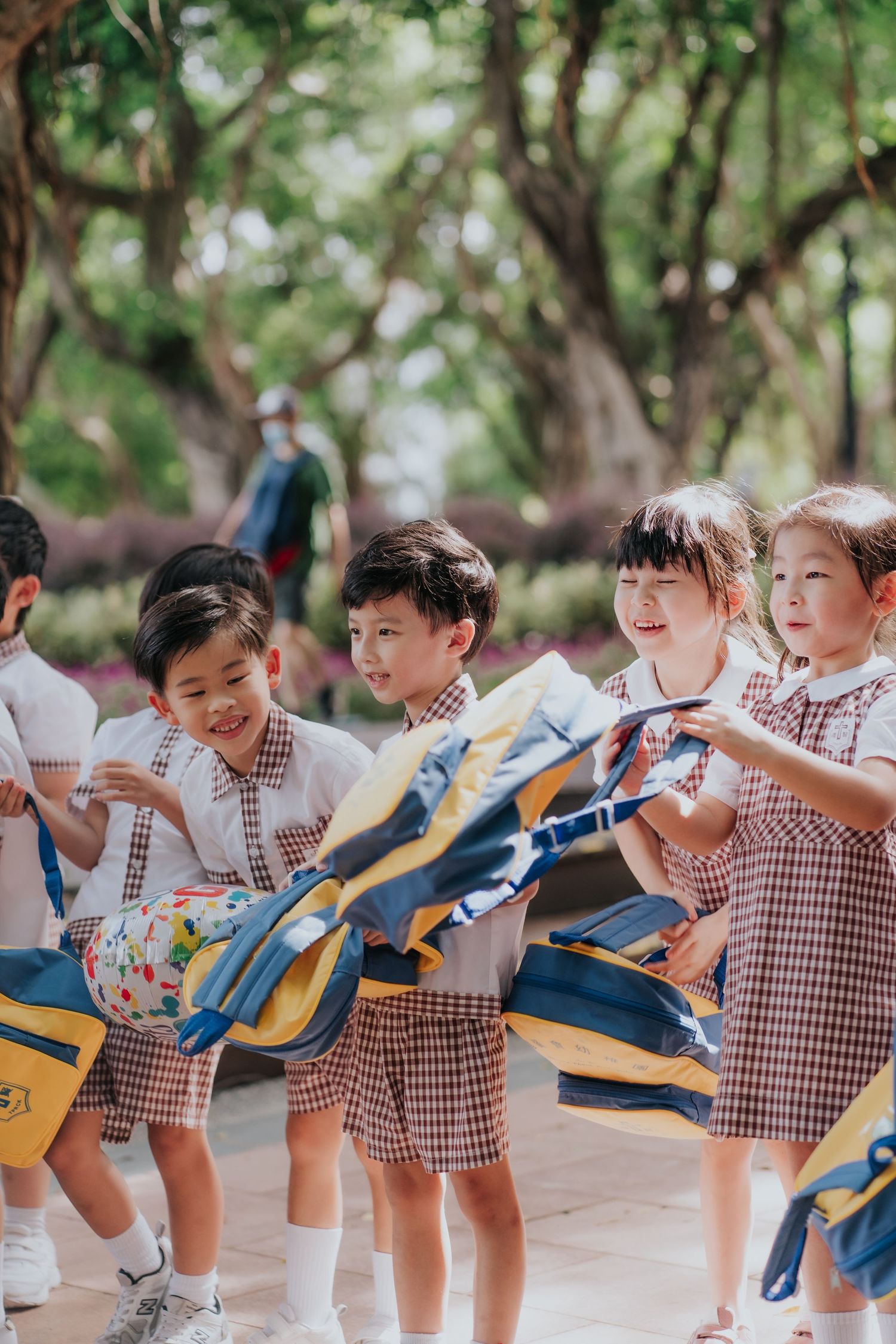 Young children in school uniforms happily holding colorful backpacks outdoors in a tree-lined park setting.一群穿著校服的小朋友背著色彩鮮豔的書包，在綠樹成蔭的公園裡快樂玩耍