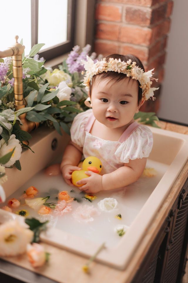 Adorable Asian baby girl in pink dress with floral crown sits in a decorative bath with a rubber duck and flowers.