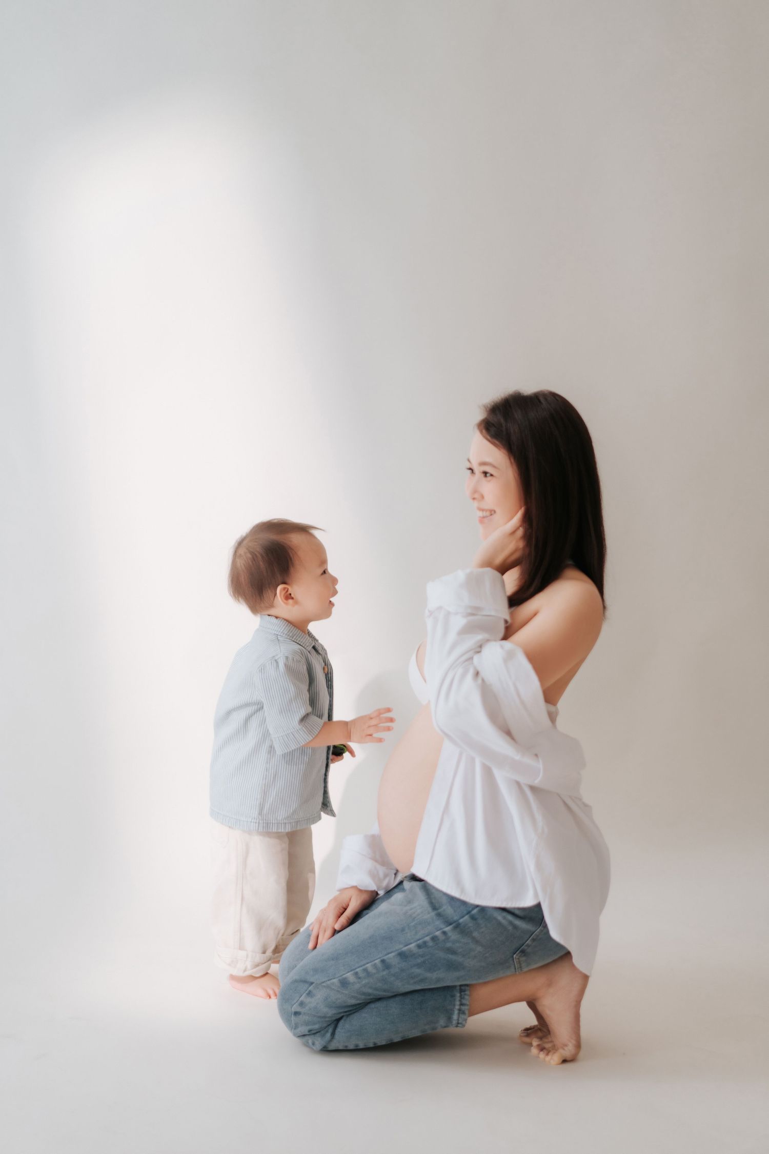 Mother and baby share playful interactions in minimalist studio setting with white background and casual attire.