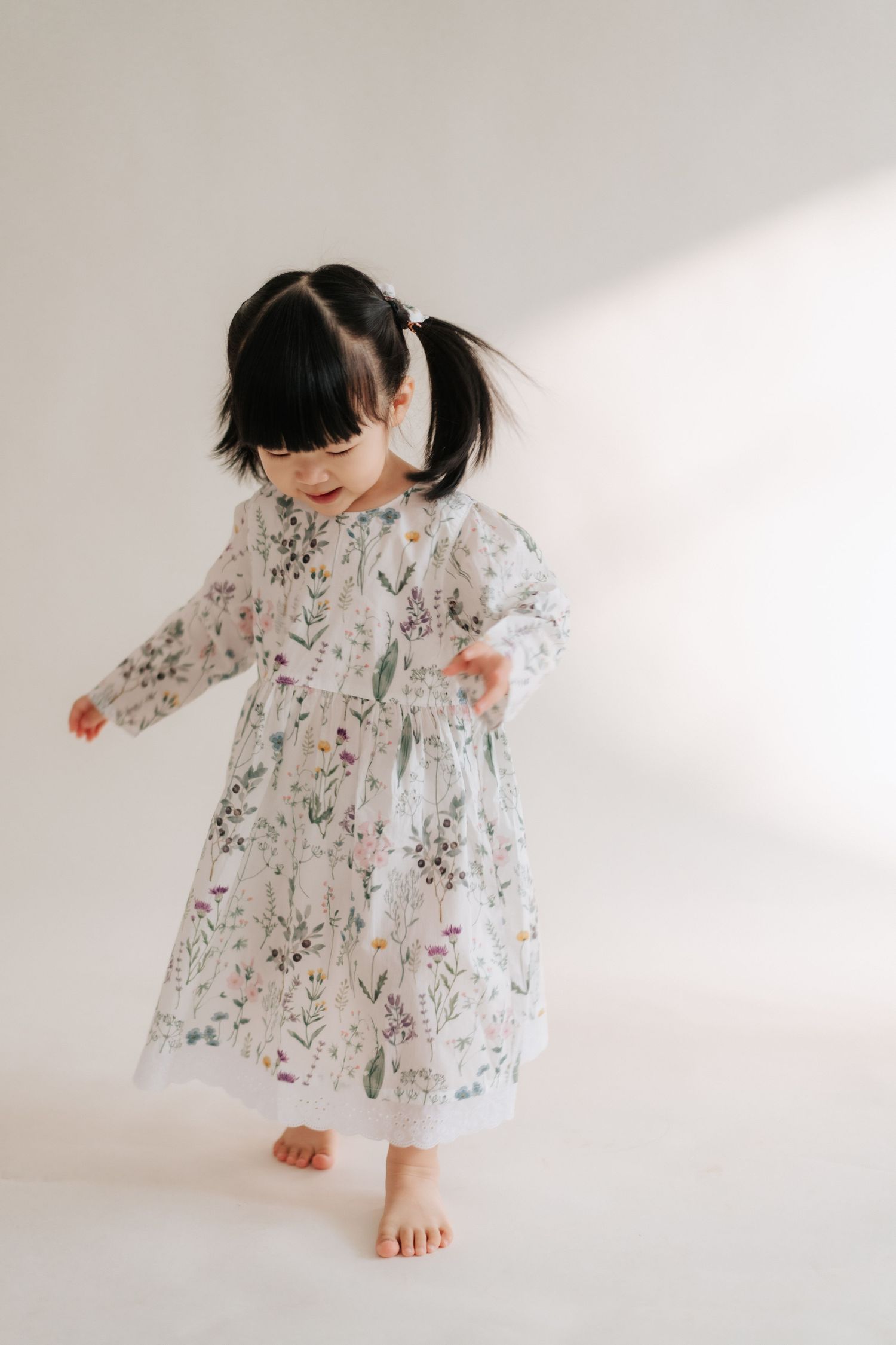 Toddler girl in a floral long-sleeve dress dancing barefoot against a white background.