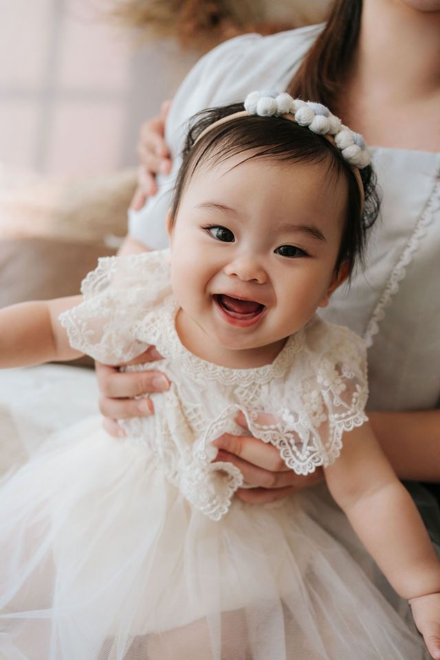 Smiling baby girl in white lace dress and floral headband held by mother during portrait session.