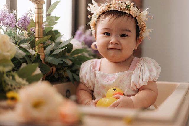 Adorable Asian baby girl wearing a floral crown smiles while holding a rubber duck among flowers.