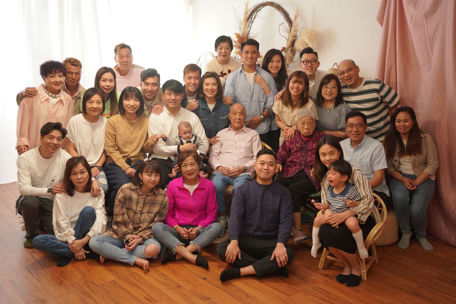 A large group of people sitting together in a row against a beige wall for a family gathering photo一大群人並排坐在米色牆邊，拍攝家庭聚會照片。