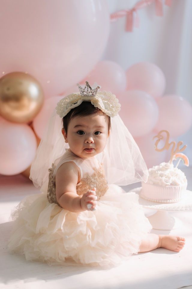 Adorable baby girl in a tulle dress and crown celebrates her first birthday with a cake and pink balloons.