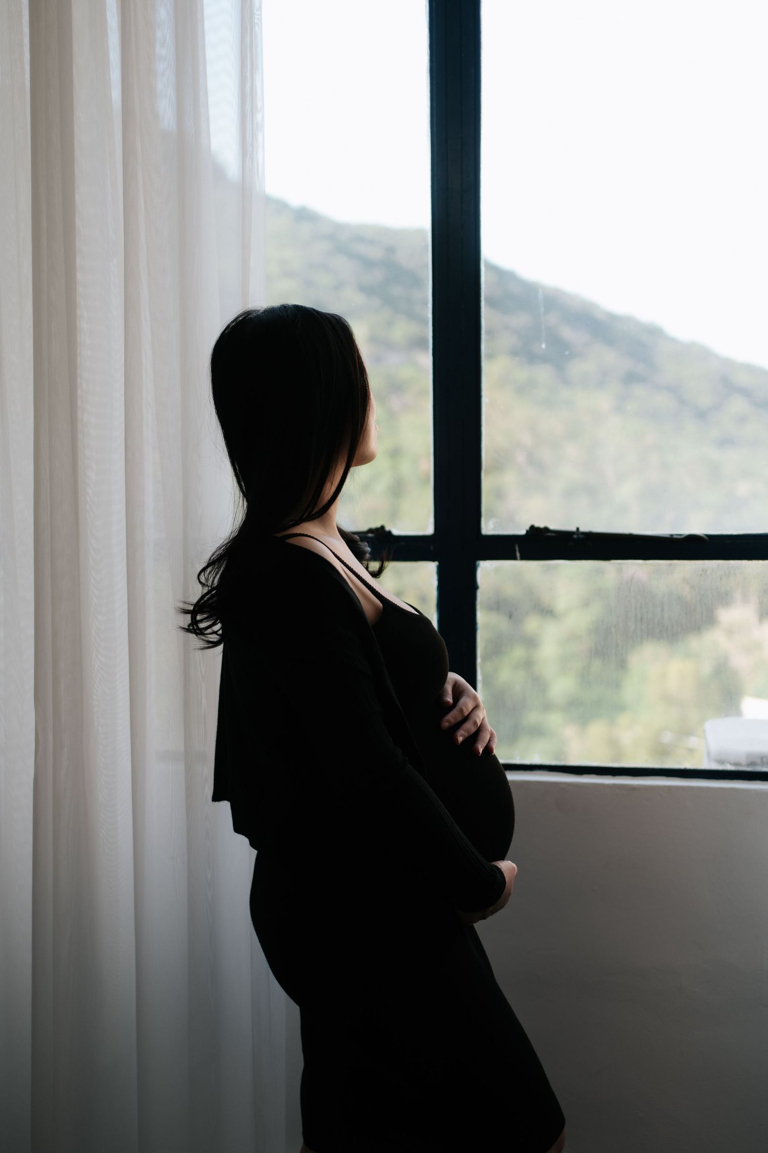Silhouette of a person standing by a window with white curtains, looking out at green mountains in soft daylight.