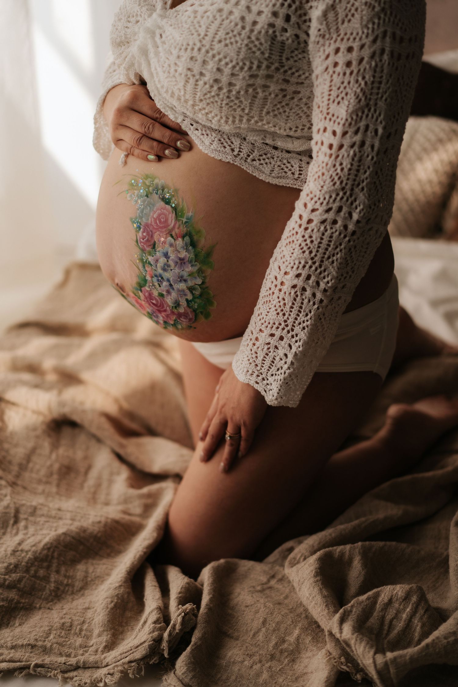Pregnant woman in white lace top sitting on linen bed, showing baby bump with colorful floral body paint design.穿白色蕾絲上衣的孕婦坐在亞麻床上，展示彩色花卉身體彩繪