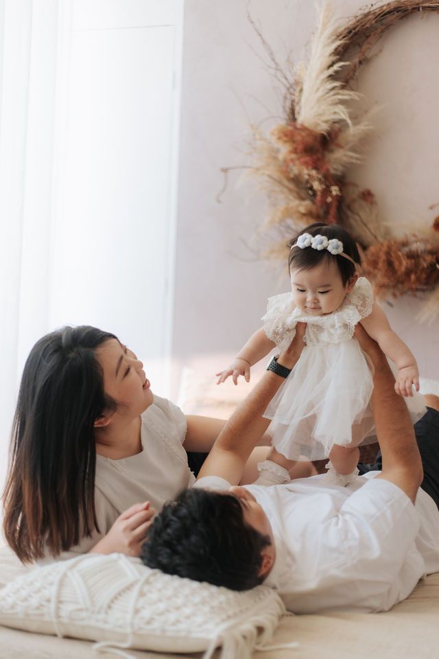 Young Asian family with mother, father, and baby girl in white dress enjoying a playful moment together on a light-colored bed.