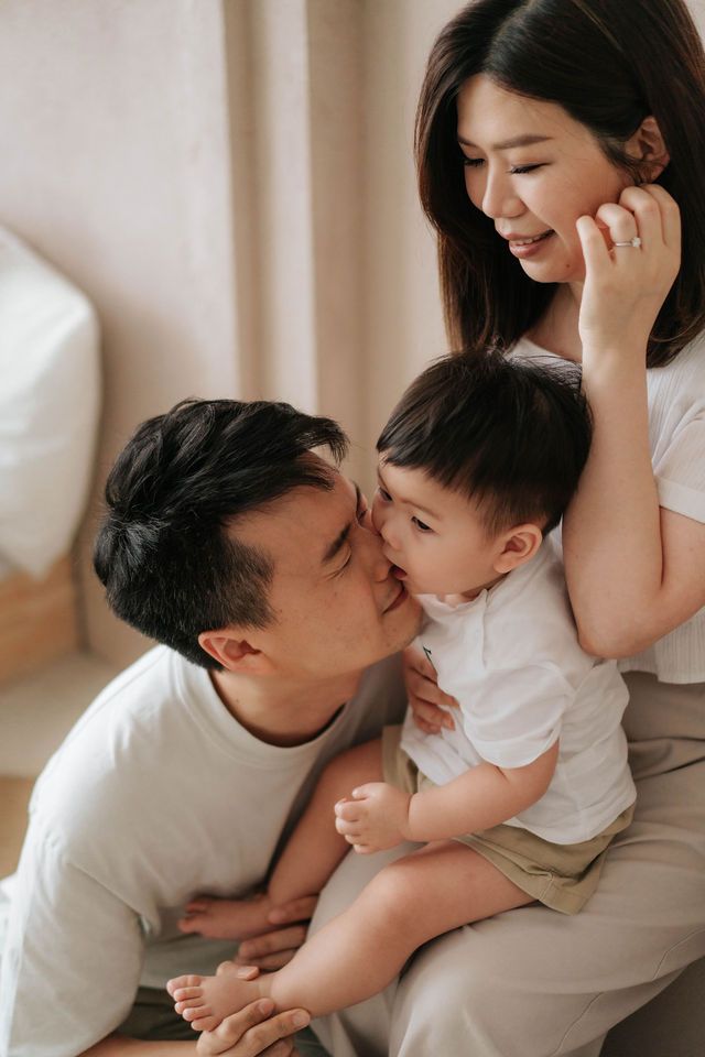 Happy Asian family with parents kissing and hugging their baby boy in a bright, cozy home setting.