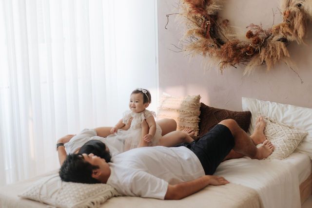 Family with baby girl relaxing on white bed near curtained window with boho wreath decor.