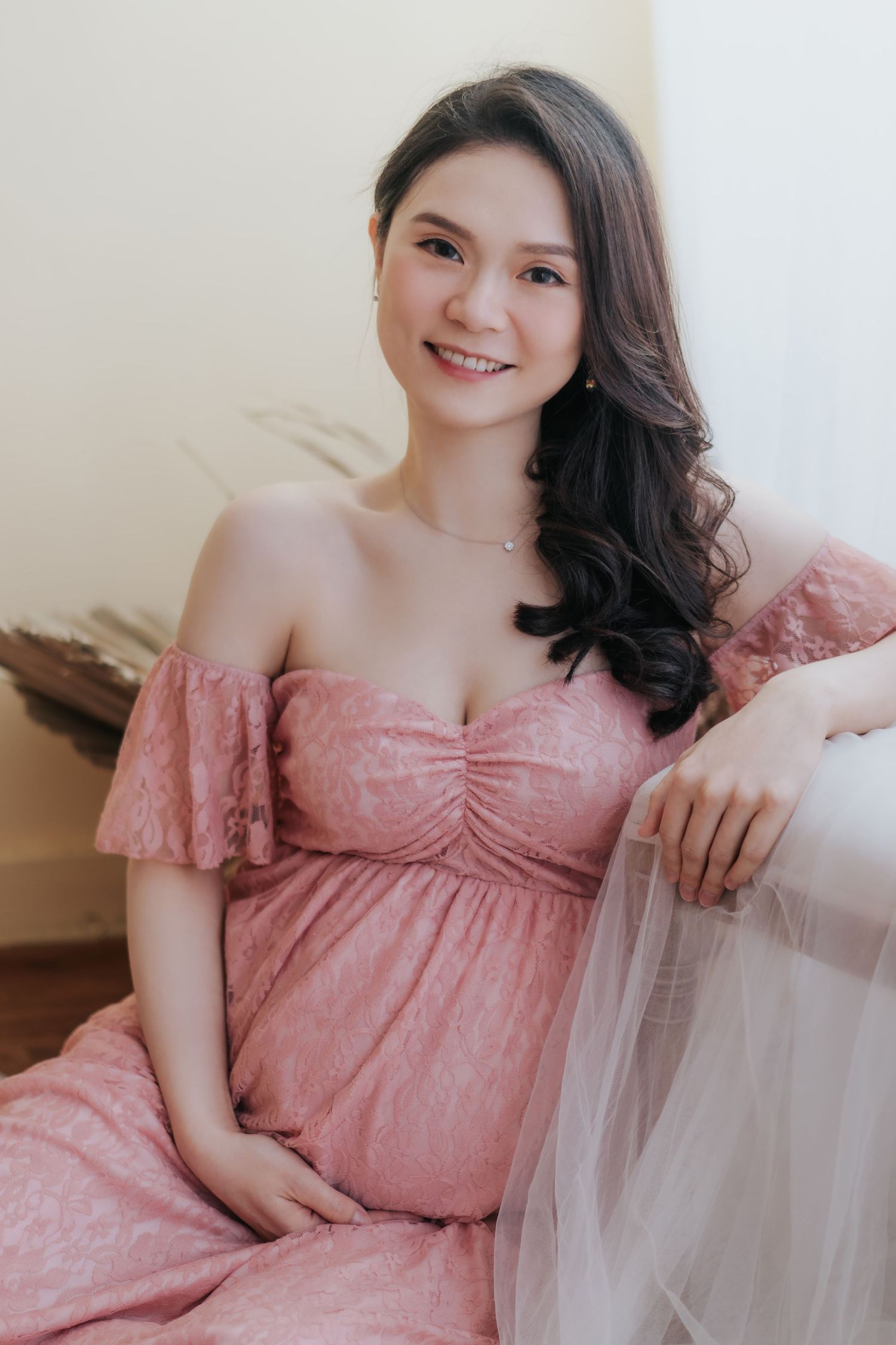 A soft and romantic maternity photoshoot featuring a pink off-shoulder dress against a light background.