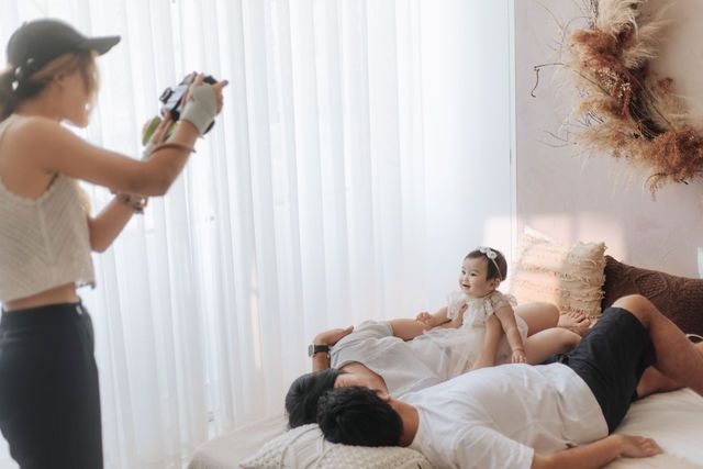 Family photo session with photographer capturing parents relaxing on bed with baby in bright white bedroom.