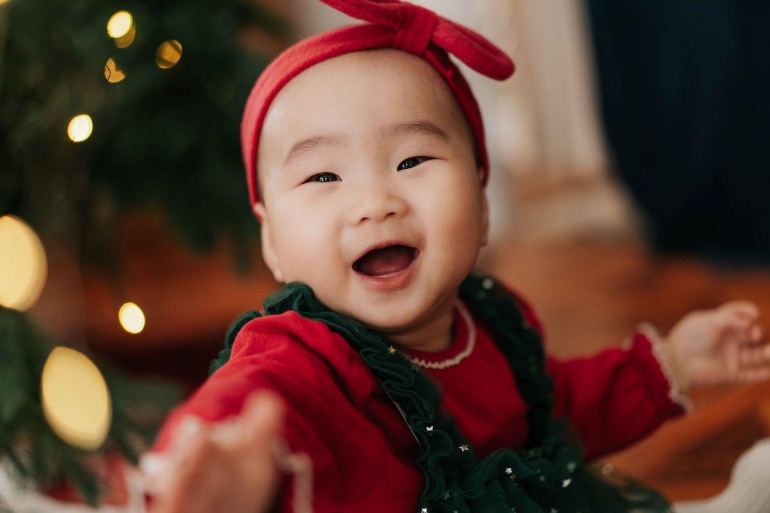 Joyful Asian baby girl in red Christmas outfit and bow headband smiling brightly near a decorated tree.